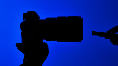Silhouette of a hand carefully holds a brush near a camera lens, against a vibrant blue background. Concept of cleaning and caring for photographic equipment.の写真素材