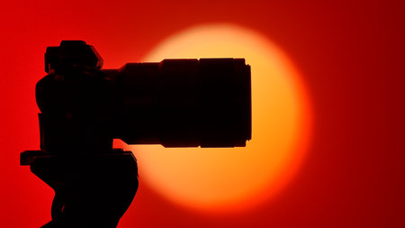 Silhouette of a professional camera with a lens mounted on a tripod on a bright round light source and a red background, creating a beautiful gradient.の写真素材
