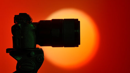 Silhouette of a professional camera with a lens mounted on a tripod on a bright round light source and a red background, creating a beautiful gradient.の写真素材