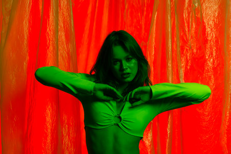 Portrait of a young female model in a green top, posing against a red background illuminated by green light with transparent plastic curtains. Color beauty, fashion art.の写真素材