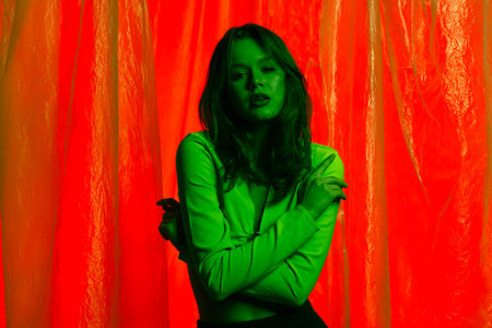 Portrait of a young female model in a green top, posing against a red background illuminated by green light with transparent plastic curtains. Color beauty, fashion art.の写真素材