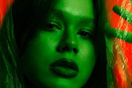 Portrait of a young female model, posing against a red background illuminated by green light with transparent plastic curtains. Color beauty, fashion art.の写真素材