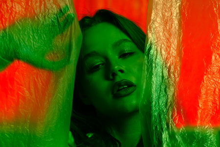 Portrait of a young female model, posing against a red background illuminated by green light with transparent plastic curtains. Color beauty, fashion art.の写真素材