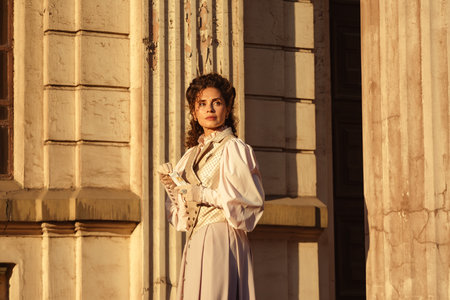 A woman in an antique dress holds a letter close to her heart near the columns of an old cathedral.