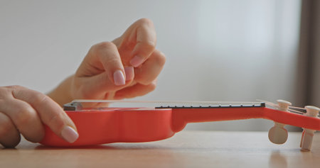 Close up of a hand playing the strings of a red toy ukulele on a tableの写真素材