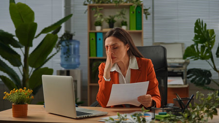 Older adult businesswoman works at green office with plants, woman sitting at desk working on laptop, checking looking at paper documents, confused stressed expression.の写真素材