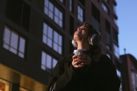 A young woman holds a coffee cup and listens to music with headphones in a modern city setting...