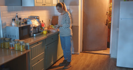 A young woman in casual clothes peels potatoes with a knife in a bright, modern kitchen in the...
