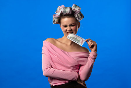 A woman in a pink blouse, with curlers shaped like banknotes in her hair against a blue background,...