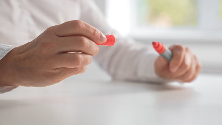 Close up of a person holding a red marker and cap over a white table, ready for writing or drawing.