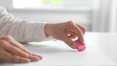 A close up of a hand using a pink eraser to remove a pencil-drawn circle from white paper.