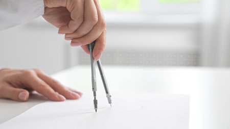 A hand skillfully uses a compass to draw precise circles on a sheet of white paper in a bright...