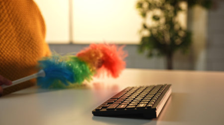 A vibrant feather duster is used to clean a computer keyboard on a bright, minimalist white desk.の写真素材