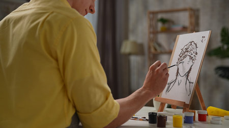 Daily lifestyle concept. Young adult man spending time at home, man in apron sits at the table and painting picture of woman portrait on portable easel, close up shot.の写真素材