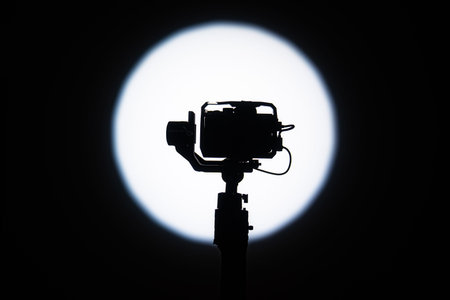 Silhouette of a video camera mounted on a gimbal stand, framed against a large circular white...