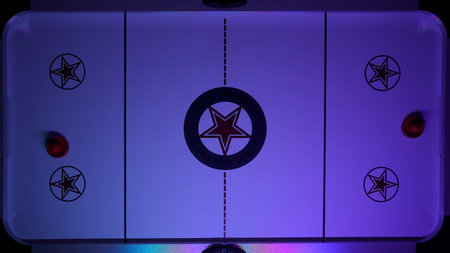 Overhead view of an air hockey table with two red strikers on opposite sides, illuminated by neon...