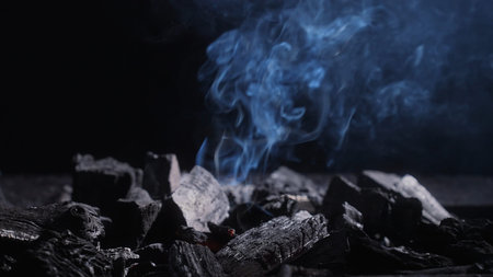 Swirling smoke above black charcoal in a grill, creating a moody background for barbecue, heat,...