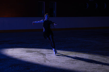 Female figure skater performing on an indoor ice rink under blue stage lighting and spotlight. Dramatic winter sports scene with elegant pose, long shadow and theatrical atmosphere.の写真素材