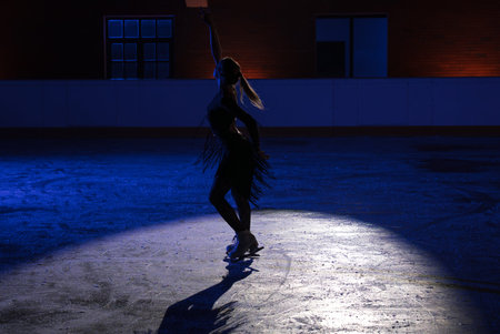 Female figure skater performing on an indoor ice rink under blue stage lighting and spotlight. Dramatic winter sports scene with elegant pose, long shadow and theatrical atmosphere.の写真素材
