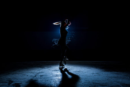Silhouette of a figure skater performing on a dark indoor ice rink with bright blue backlight and...