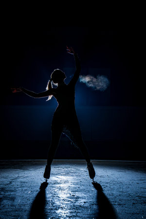 Silhouette of a figure skater performing on a dark indoor ice rink with bright blue backlight and...