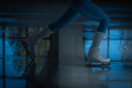 Close up of figure skater legs and blades on a shiny indoor ice rink with reflection and shadow....