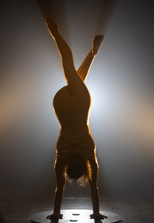 Full shot silhouette of a young circus gymnast performs a arms handstand in a dark studio against a...