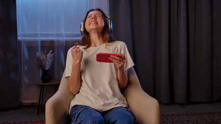 A young woman with headphones sits indoors, joyfully celebrating as she looks at her smartphone,...