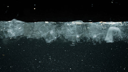 Close up of ice layer floating on water with oxygen bubbles and particles in dark background....