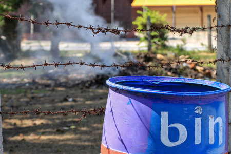A blue trash can sits outside next to a barbed wire fence, with smoke from burning garbage rising from behind.の写真素材