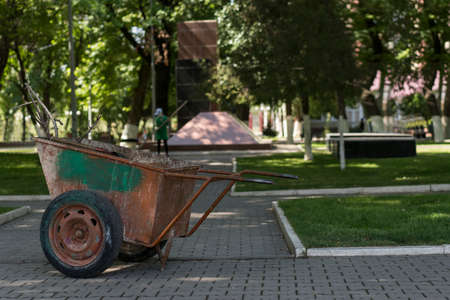 In the background out of focus, a janitor is manually sweeping a walkway in a park. In the foreground a shabby metal wheelbarrow for garbage disposal.の写真素材
