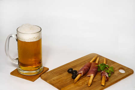 Glass mug of beer on a wooden board. Foam and drops. Breadsticks wrapped in ham with olives and a sprig of parsley.の写真素材