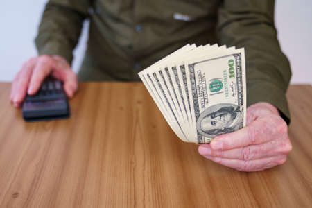 A man's hand holds out one hundred dollar banknotes. Calculator in the right hand.