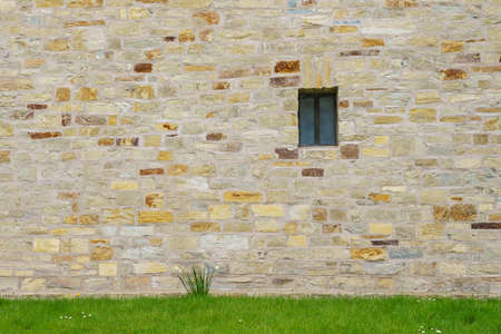 An ancient stone wall of hewn stone with a small window. Green grass in the foreground.の写真素材