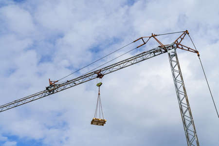 A wooden euro pallet lifted by a tower crane against a blue sky with light clouds.