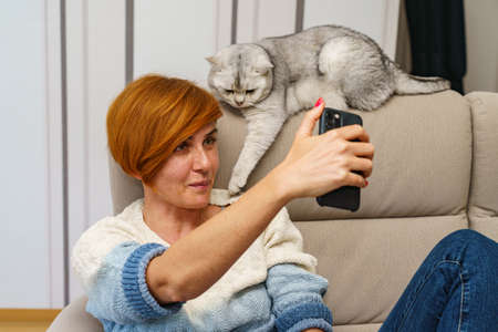 A girl with cute freckles sits on the couch and takes a selfie with a British shorthair cat lying on...