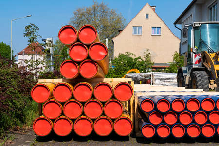 Construction site in city. Stack of orange-yellow and blue PVC conduit pipes. Tractor next to.