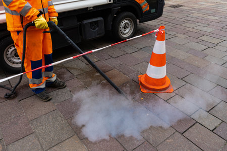 Road worker cleans the pavement with a steam pressure hose. Orange-white conical pylon of fencing tape.の写真素材