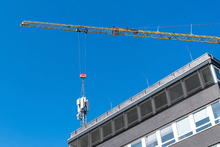 Installation of a 5G broadcast tower on the roof of a building using a tower crane. Against the backdrop of a clear blue sky.の写真素材