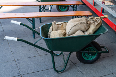 Green wheelbarrow with sacks stands near wooden benches.の写真素材