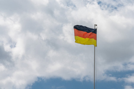 German national flag on a metal mast flutters in the wind against a cloudy sky.の写真素材