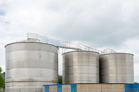 Three large silver metal tanks against a cloudy sky. A metal staircase with railings passes from above.の写真素材