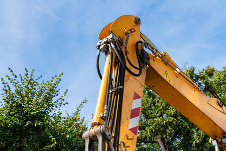 Part of the boom of an orange excavator with a hydraulic drive against a background of blue sky and green tree branches.の写真素材