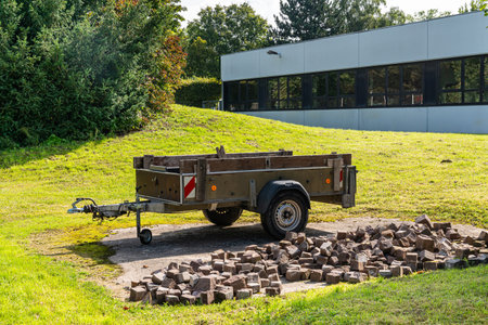 Trailer for a car next to a pile of cobblestones for paving the sidewalk. Trees and buildings in the...