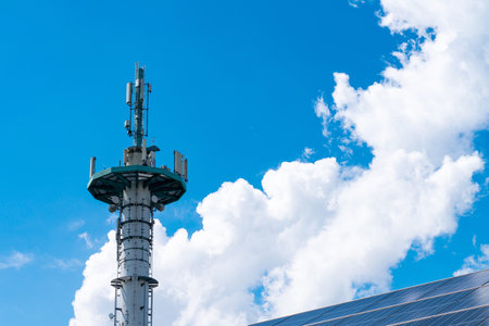 Broadcast mast and part of the roof with solar panels against a blue sky with white clouds.の写真素材