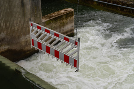 A protective fence on the side of an open dam with a white and red striped barrier, surrounded by flowing water.の写真素材