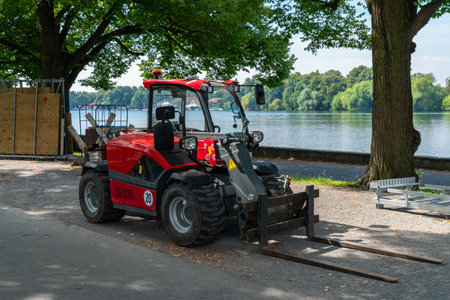 A red compact loader is parked by the riverbank, surrounded by green trees, enjoying a bright sunny...