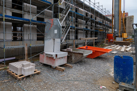 Construction materials and machinery are arranged at a building site with scaffolding and gravel...