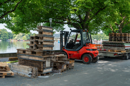 A forklift transports wooden pallets beside a calm lake, surrounded by greenery and outdoor activity.の写真素材