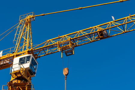 A large yellow construction crane stands tall against a clear blue sky. Its operator's cabin is visible, along with a hook dangling down, ready for heavy lifting at a building site.の写真素材